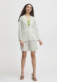 Hellbeige Leinenblazer mit zwei vorderen Taschen, kombiniert mit passenden Shorts mit Fransenabschlüssen. Darunter ein grünes geripptes Tanktop.