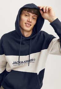 Sudadera en azul marino y crema con cuerda en la capucha, bolsillo frontal y texto del logo en azul oscuro. Tejido suave con un ajuste deportivo y relajado.