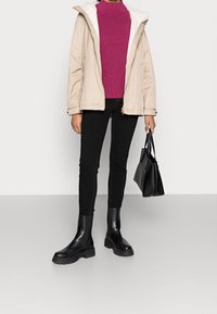Beige middellange jas met witte fleece voering, gecombineerd met een magenta ribbeltrui, zwarte skinny jeans en enkellaarsjes in zwart. Zwarte handtas.