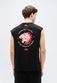 Μαύρο αμάνικο μπλουζάκι με μεγάλη εικόνα ενός βρυχώμενου λευκού και κόκκινου τίγρη στην πλάτη, με έντονα γράμματα και ένα λογότυπο στο κάτω κέντρο.