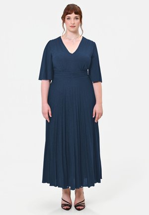Donna con pelle chiara e capelli rossi che indossa un lungo vestito blu scuro con maniche corte e tacchi neri con cinturini, in piedi su sfondo bianco.