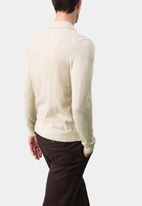 Gebreide trui in crème met lange mouwen en een polo-kraag, voorzien van een geribbelde zoom en boorden, gecombineerd met donkere bruine broek.