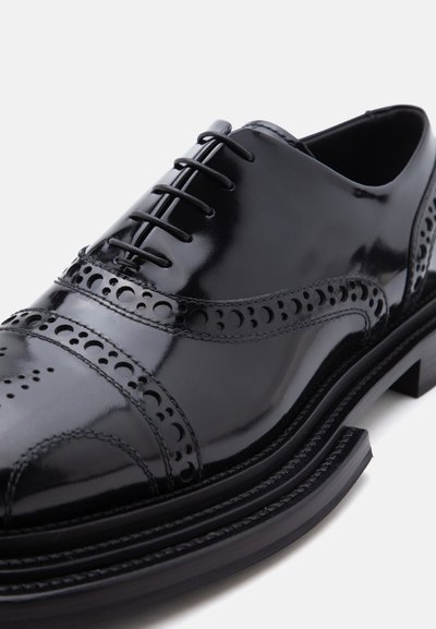 Μαύρο γυαλιστερό δερμάτινο παπούτσι Oxford με διακοσμητικές τρύπες brogue και κλειστό κορδόνι σε λευκό φόντο.
