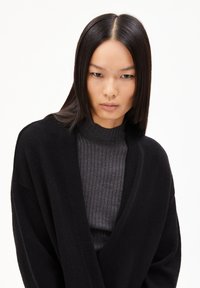 Schwarze Strickjacke mit lockerer Passform und weiten Ärmeln, getragen über einem dunkelgrauen gerippten Rollkragenpullover mit glatter Textur und minimalistischem Design.