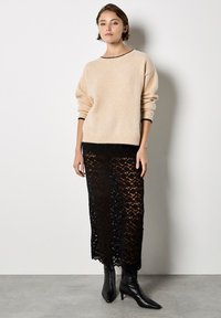 Suéter de punto beige con mangas largas y un escote negro, combinado con una falda negra de encaje transparente y botas negras de punta.