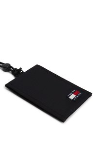 Μαύρο υφασμάτινο πορτοφόλι σε παραλληλόγραμμο σχήμα με ένα πατς λογότυπου Tommy Jeans και μαύρο κορδόνι κρεμαστής θήκης προσαρτημένο σε μία γωνία.