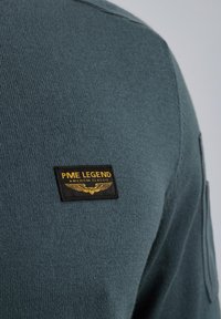 Dunkelblaues Strickpullover mit einem schwarzen und goldenen Stoffetikett, auf dem der Text "PME LEGEND AMERICAN CLASSIC" steht. Glatte Textur, lässiger Stil.
