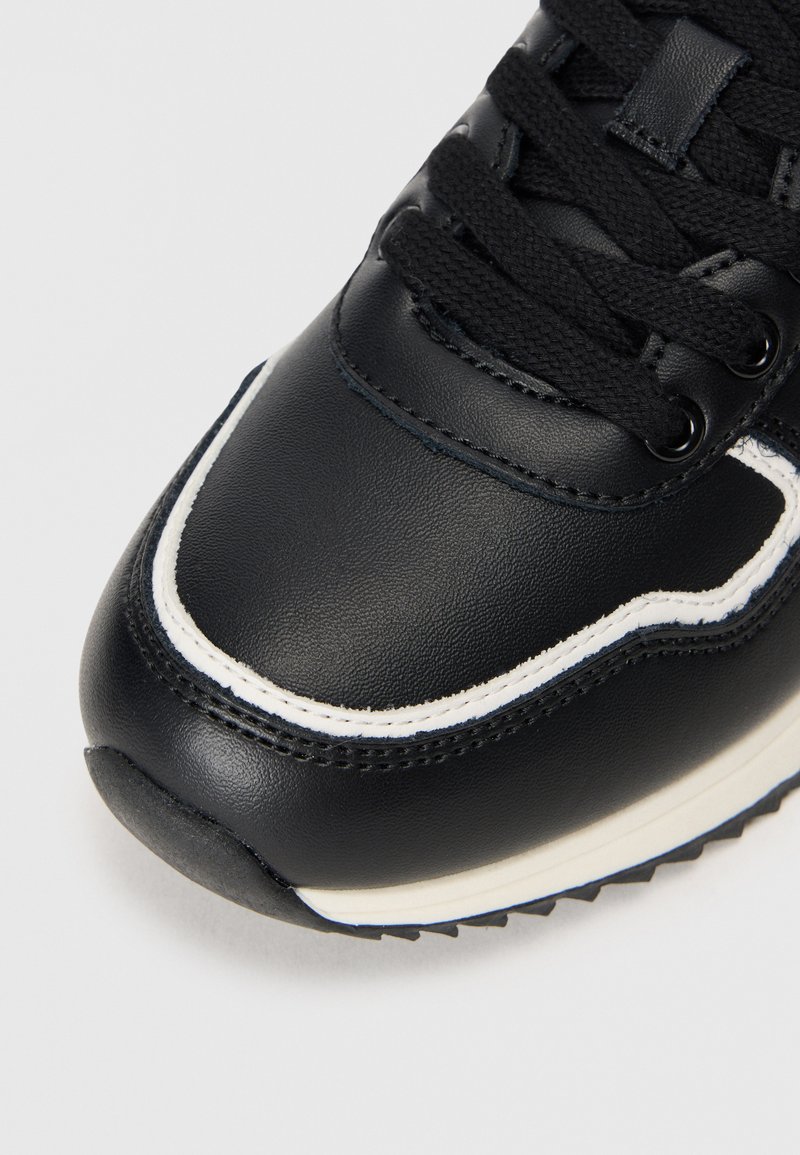 Zapatilla negra con parte superior de cuero liso, costuras blancas, diseño con cordones y suela de goma texturizada con un patrón estriado.