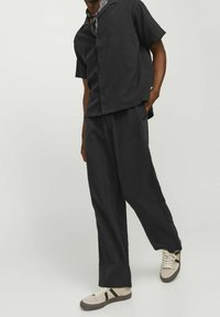 Mies, jolla on yllään löysä musta lyhythihainen nappipaita, mustat leveälahkeiset housut, beiget lenkkarit ja kuvioitu kaulahuivi.