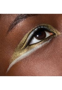 Gros plan d'un œil mettant en valeur un eyeliner doré et blanc scintillant sur une peau foncée, avec une texture de peau détaillée et des cils bien définis.