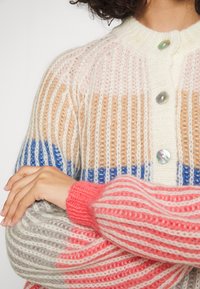 Pruhovaný pletený kardigan s kombinací krémové, béžové, růžové, modré a korálové barvy. S knoflíky na přední straně s irizujícími knoflíky a žebrovanými manžetami.