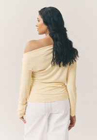 Haut à manches longues et épaules dénudées en tissu jaune doux, coupe ajustée, avec poignets côtelés, assorti à un jean blanc, modèle vu de dos.
