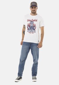 T-shirt blanche avec imprimé graphique rouge et bleu d'un moteur rétro et des flammes, portée avec un jean bleu et des baskets noires. Casquette marron avec logo.