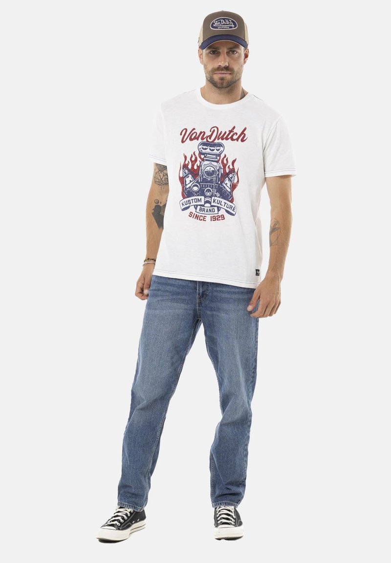 T-shirt blanche avec imprimé graphique rouge et bleu d'un moteur rétro et des flammes, portée avec un jean bleu et des baskets noires. Casquette marron avec logo.