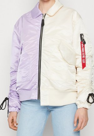 Puoliksi violetti, puoliksi kerma valkoinen vetoketjullinen bomber-takki taskuilla, mustat nyörit hihoissa ja punainen "Remove Before Flight" -merkki vasemmassa hihassa.