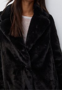 Zwarte nep bontjas met een grote, omgeslagen kraag. De textuur is zacht en pluche, met een dubbelgeknoopte sluiting en een subtiele glans.