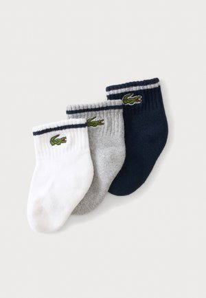 Tre babystrømper i hvid, grå og navy blå, hver med en sort stribe nær kanten og et grønt krokodillelogo på siden.