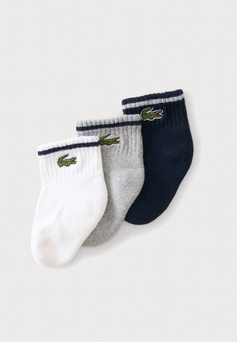 Trois chaussettes pour bébé en blanc, gris et bleu marine, chacune avec une bande noire près du bord-côte et un logo crocodile vert sur le côté.