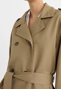 Khaki trench coat with double-breasted buttons, large notched collar, and a fabric belt at the waist. Smooth texture, tailored fit.