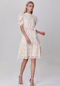 Robe en dentelle crème avec des manches bouffantes, un devant à boutons et une ceinture à nouer. Présente une jupe à volants avec un ourlet festonné et un motif floral.