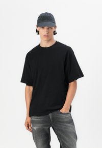 Tricou din bumbac negru, cu guler rotund și mâneci scurte, asociat cu blugi gri deteriorați și o șapcă gri cu un design brodat.