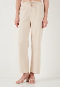 Beige, relaxed-fit pants with an elastic waistband and drawstring. Smooth texture; wide-leg design. Paired with strappy light-colored sandals.