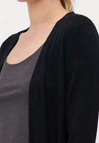 Zwarte cardigan van zacht materiaal, met een open voorkant en een gladde textuur. Gedragen over een grijze tanktop.