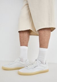 Sneakers bianchi in pelle con suole in gomma, caratterizzati da dettagli perforati, lacci e un logo sulla linguetta. Indossati con calzini bianchi e pantaloni corti beige.