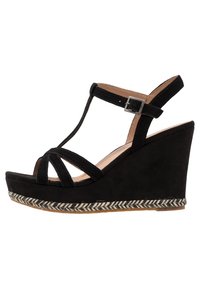 Czarne zamszowe sandały na koturnie charakteryzują się zamkniętym noskiem, designem w stylu T-strap, plecionym akcentem z juty wokół podstawy oraz regulowanym zapięciem na kostce.