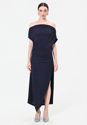 Femme portant une robe sombre épaules dénudées avec des paillettes, une fente haute et des talons hauts argentés, debout devant un fond blanc uni.