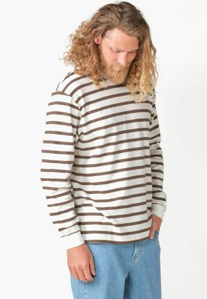 Vousatý muž s dlouhými kudrnatými vlasy, který nosí bílou košili s hnědými vodorovnými pruhy a světlé modré džíny, dívá se dolů.