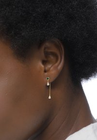 Goldene Ohrringe mit einem Smaragd-Akzent und zwei hängenden, klaren, birnenförmigen Steinen an einer zarten Kette. Glatte Textur und elegantes Design.