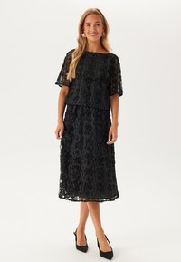 Svart spetsklänning med blommönster, korta ärmar och knälång kjol. Strukturerat tyg med lagerdesign och en avslappnad passform.