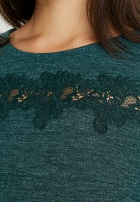 Zielono-turkusowy top z koronkowymi detalami i skomplikowanymi wzorami wzdłuż dekoltu, wykonany z miękkiej, teksturowanej tkaniny.