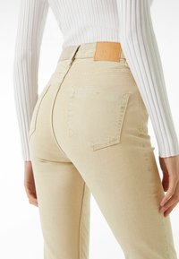 Beiga högmidjade jeans med slät yta, två bakfickor och en brun läderetikett på midjan. Ribbade vita toppen synlig delvis.