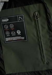 Interno della giacca da performance verde con tessuto idrorepellente e antivento. Presenta una tasca con cerniera e un'etichetta di cura con icone delle proprietà.