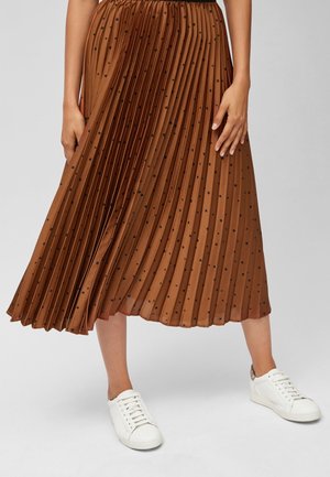 Brązowa plisowana spódnica midi w czarne kropki, noszona z białymi trampkami, ukazująca nogi i ręce na jednolitym tle.