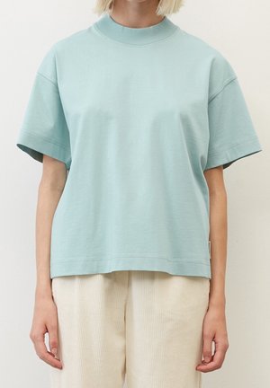 Frau trägt ein hellblaues, übergroßes T-Shirt mit kurzen Ärmeln und beige Cord-Hosen, steht vor einem schlichten Hintergrund.