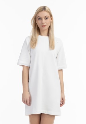 Młoda kobieta z długimi blond włosami, ubrana w prostą białą sukienkę z krótkim rękawem, stoi na białym tle.
