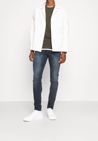 Veste blanche avec boutons, pull vert, jean bleu foncé, et baskets blanches. Design simple, coupe d écontractée, et texture lisse partout.