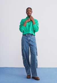 Sweatshirt verde cropped com meia abertura, combinado com calças cargo de cor azul clara, apresentando vários bolsos e um corte solto. Sapatos castanhos.