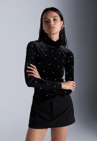 Svart fløyels høyhalset topp med lange ermer, dekorert med små hvite perler jevnt fordelt over stoffet; parret med et svart mini skjørt.