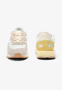 Een paar crèmekleurige en beige sneakers, gezien van voor en achter, met veters, suède details en gele accenten op de hielen.