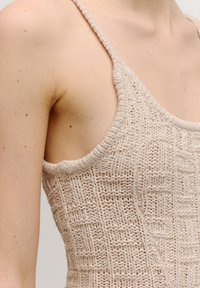 Beige gestricktes Oberteil mit dünnen Trägern, das ein strukturiertes Gittermuster und einen hohen Schnitt um den Halsausschnitt und die Armlöcher aufweist.