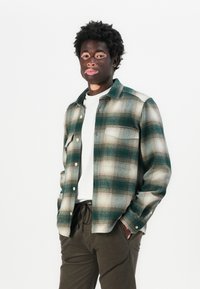 Groen en beige geruite flanellen shirt met een knoopsluiting aan de voorkant, twee borstzakken en een relaxte pasvorm, gecombineerd met donkere broeken.