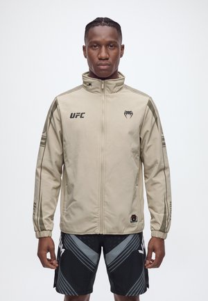 Un hombre con una chaqueta beige de UFC con cremallera y unos pantalones cortos negros estampados, de pie sobre un fondo claro y liso.