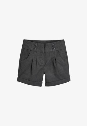 Smilende pige iført en hvid bluse med lange ærmer, grå højtaljede shorts, sorte strømpebukser og sorte støvler, stående mod en ensfarvet baggrund.