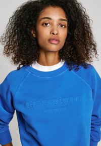 Felpa blu con colletto a girocollo rigato e maniche raglan, con testo in rilievo "PEPE JEANS LONDON" e "WEST ELEVEN" sul davanti.