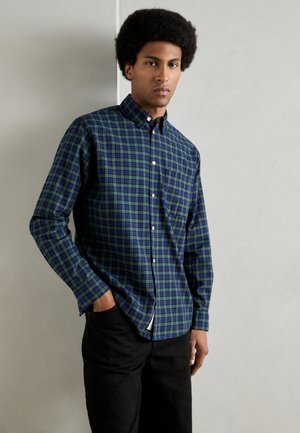 Camisa de cuadros azul y verde, con cuello, mangas largas y un único bolsillo en el pecho, combinada con pantalones negros.