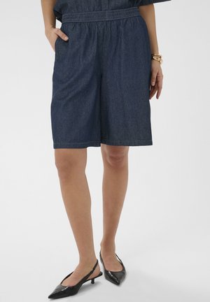 Žena oblečená v tmavomodrých riflových šortkách po kolená, s čiernymi špicatými sandálmi na opätku so zadným remienkom a zlatými náramkami na pravej ruke, stojaca s ľavou rukou v vrecku.